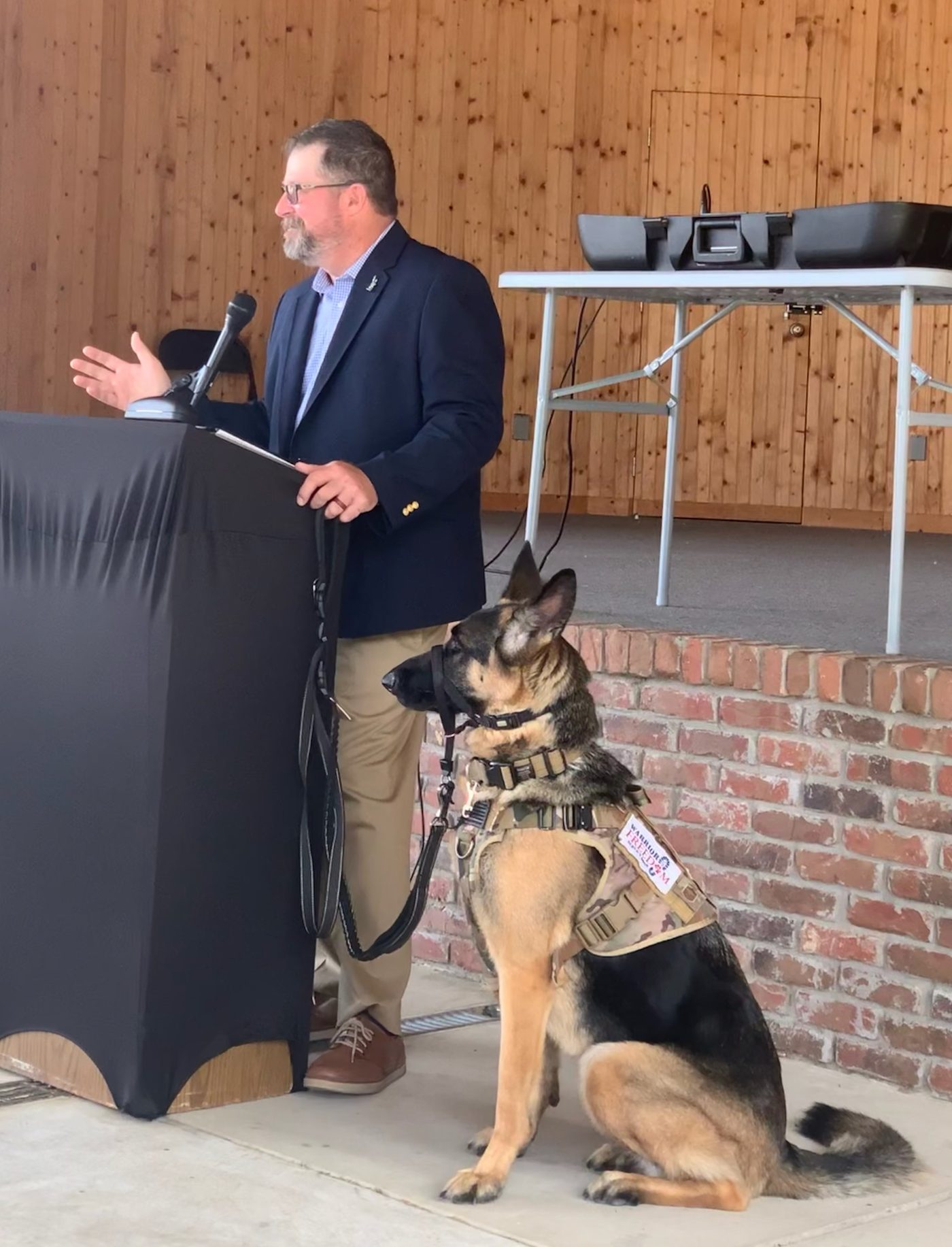 Eric Dudash speaking at a lectern with his service dog Phantom sitting beside him