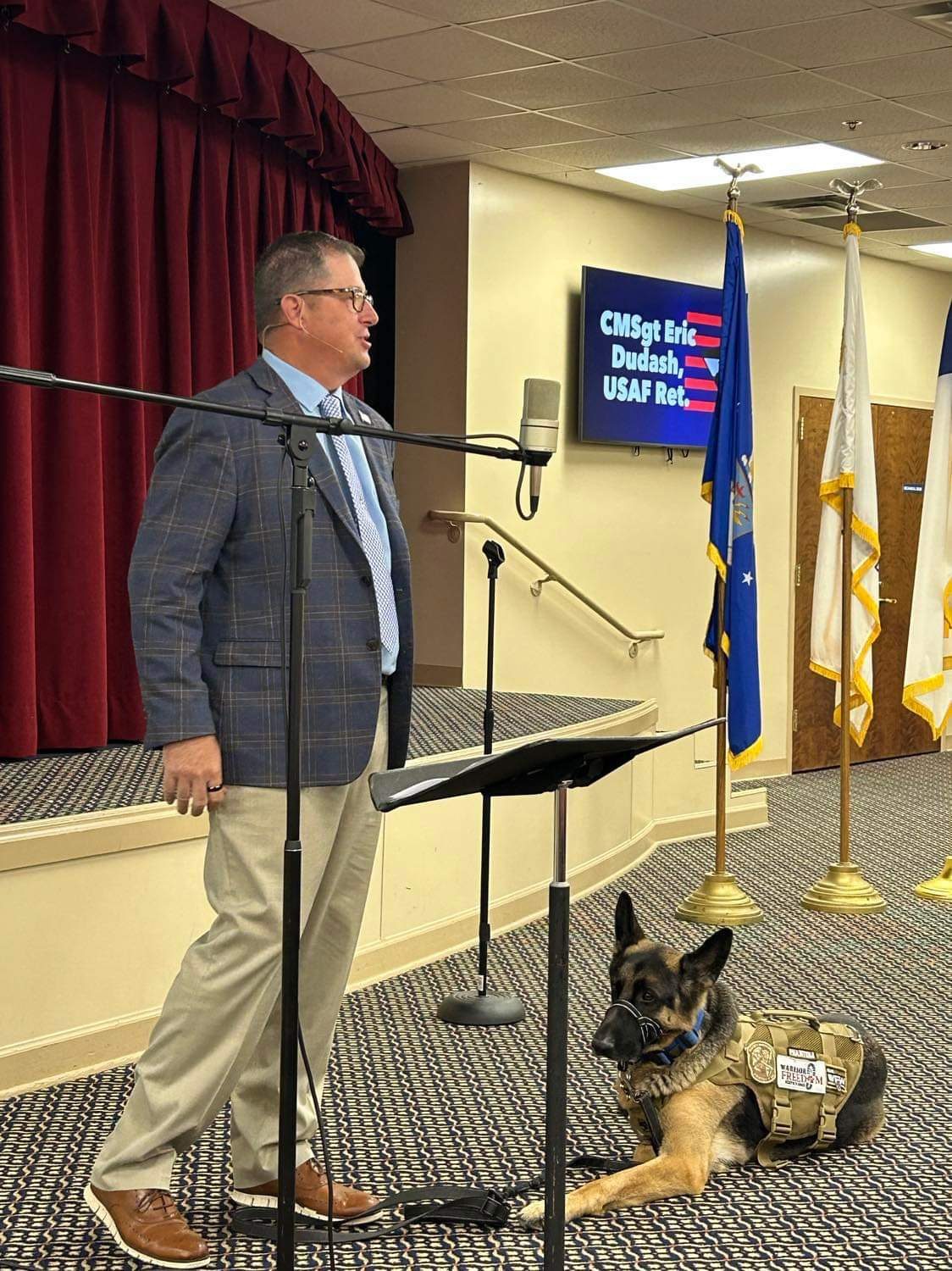 Eric Dudash at a microphone, a screen behind him reading CMSgt Eric Dudash, USAF Ret., with the Air Force flag and colours; Phantom lies at his feet in a Warrior Freedom service vest